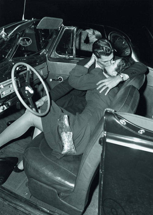 A couple kissing in the front seat of a convertible car at a drive-in movie theater, 1940s. The man in the next car ignores the couple, resting his elbow on the window.