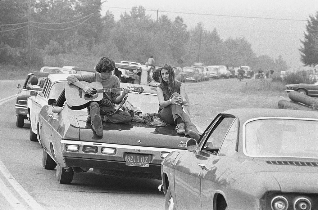 A couple play the guitar sitting on their car on the way to the Woodstock Festival, August 1969. (Photo by Baron Wolman/Iconic Images/Getty Images)
