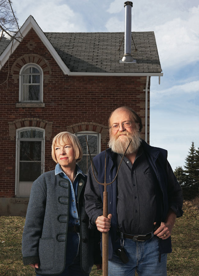 Organic farming couple posing in front of their farmhouse. The husband is holding a pitch fork, sporting a large grey beard while the wife is sporting a short blonde haircut.