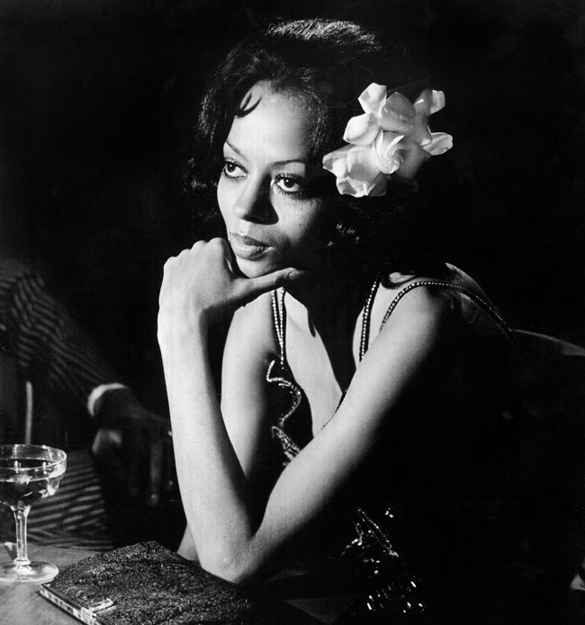 A black and white photo of Diana Ross, leaned forward resting an elbow on the table and her hand on her chin. 