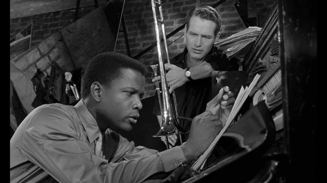 Sidney Poitier writing on a notepad at a music stand while Paul Newman, holding a trumpet, looks on. 