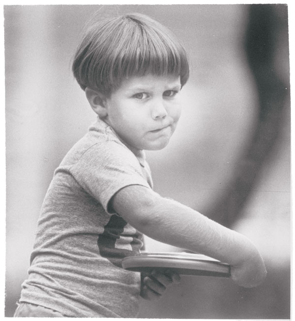 A black and whit photo of a young boy with a bowl cut throwing a frisbee.