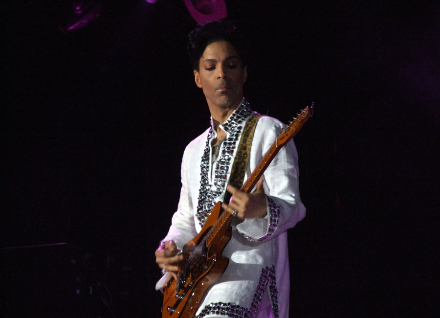 The singer Prince strums his guitar, wearing a white sports coat.