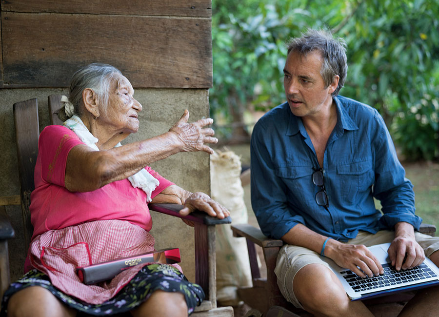 Dan Buettner converses with an older woman as he takes notes on his lap-top. 