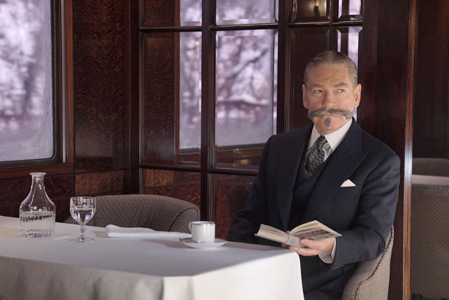 Kenneth Branagh as Hercule Poirot in Murder On The Orient Express dressed in a dark suit holding a book open with one hand. 