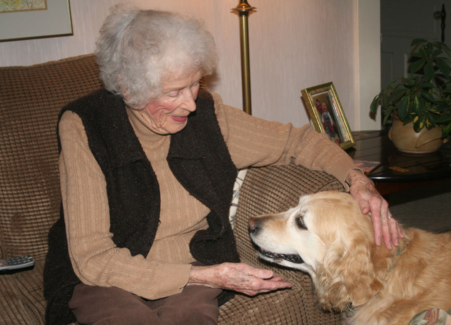 A senior woman sits on a sofa and pets her golden retriever seated at her feet