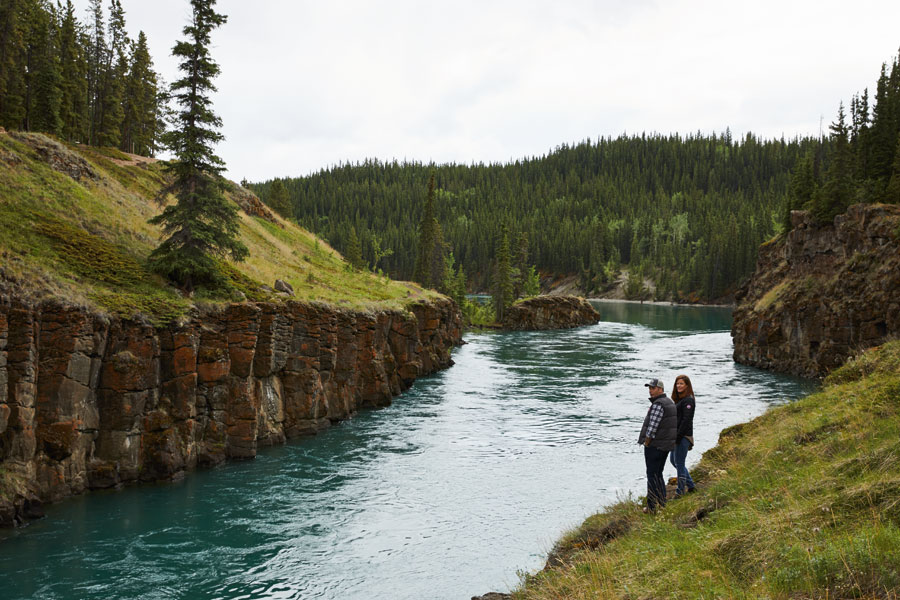 Two people stand at at the edge of turquoise water surrounded by lush grass and pine trees. 
