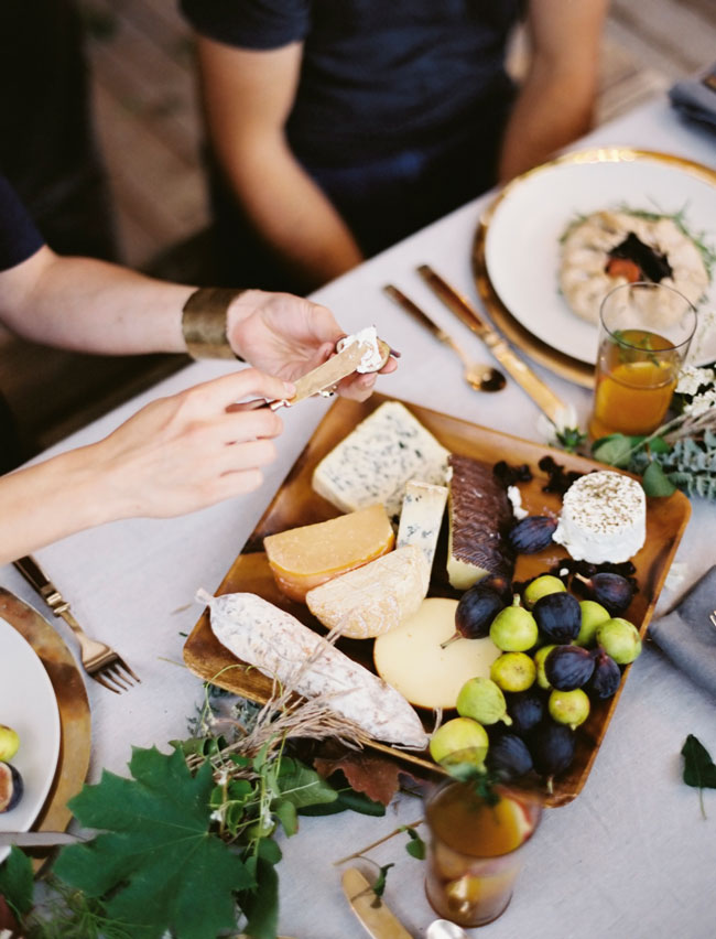 An assortment of cheese on a cheeseboard with a pair of hands spreading a soft cheese on a cracker. 