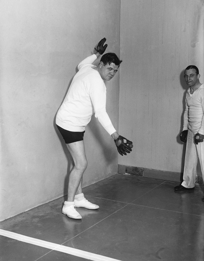 Babe Ruth holds the ball up to serve at a handball court. 