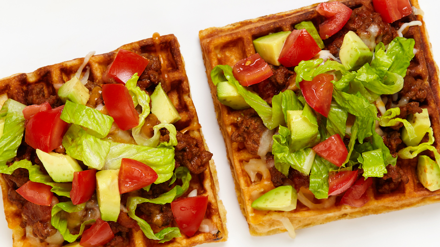 Two waffles with taco ingredients on it including ground beef, tomato and avocado. 