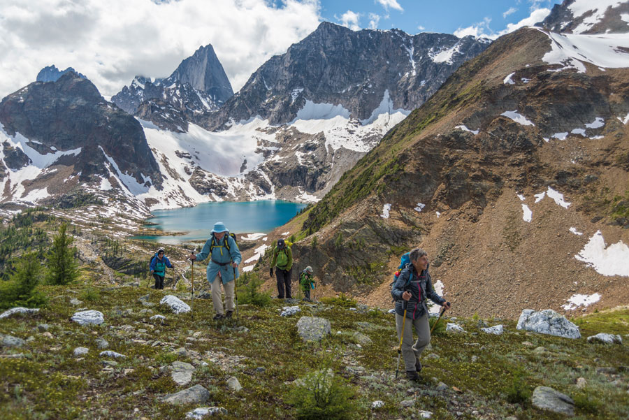 A group of people hiking a mountain. Snow covered mountains and bright blue water can be seen ahead. 