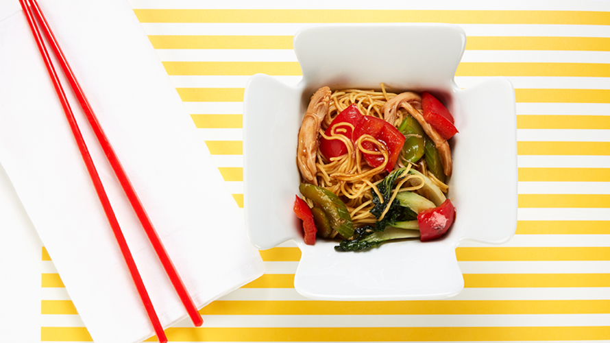 Chicken and vegetable lo mein in a Chinese takeout box with a pair of red chopsticks laying beside it. 