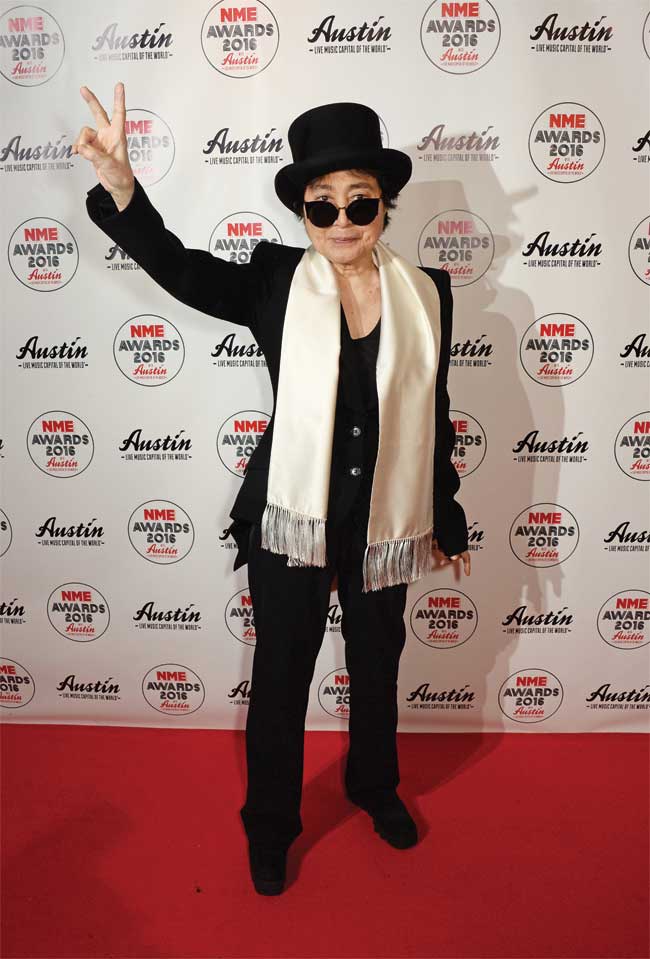 Yoko Ono throws up a piece sign at a red carpet event dressed in a black suit and a gold scarf. 