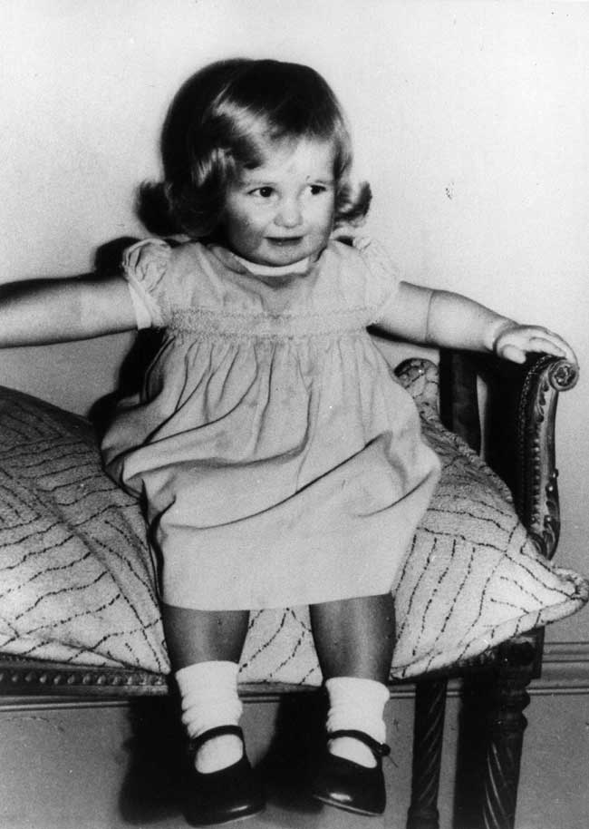 Princess Diana as a toddler sitting on a cushioned chair with her arms resting on the arm rests.