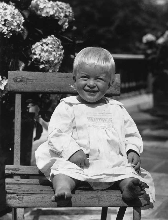 Prince Philip as an infant sitting on a wooden chair squinting and smiling.