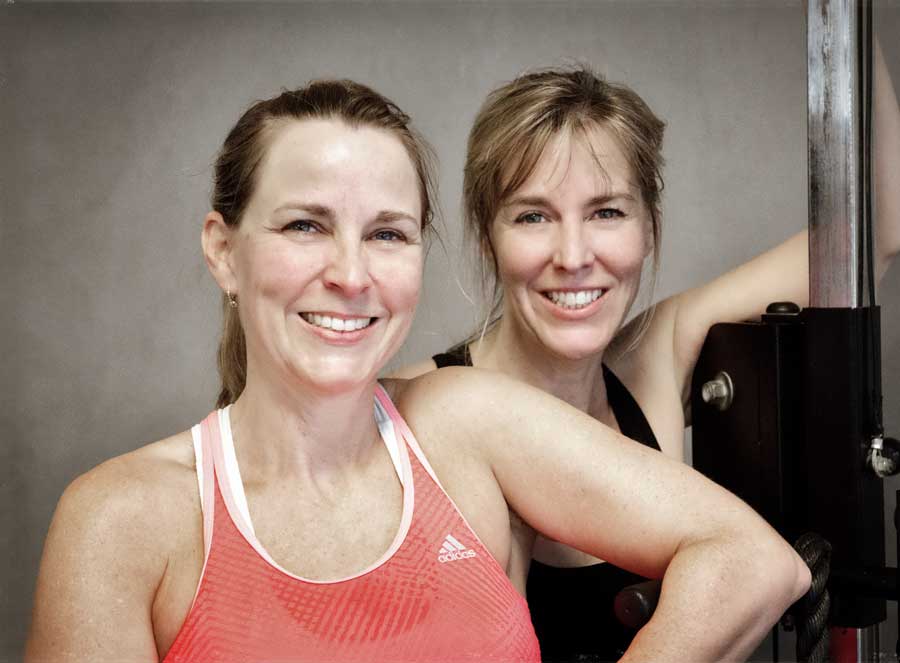 Two women posing together smiling while leaning against an exercise machine.