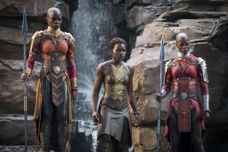 Three Wakanda warrior women stand in front of a waterfall, each wearing futuristic armour with a tribal element to it as well. 