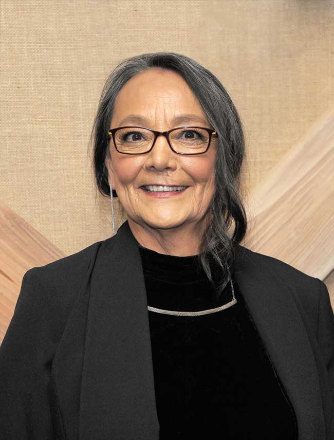 Tantoo Cardinal wearing glasses and a dangly earring, smiling. 