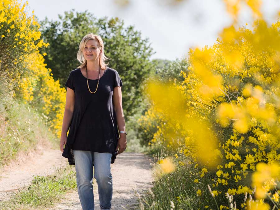Debbie Travis standing on a path smiling with yellow flowers on each side.