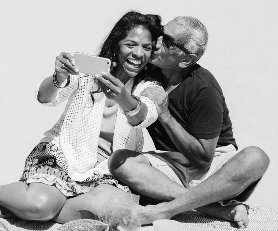 A woman taking a selfie laughing as a man kisses her on the cheek. 