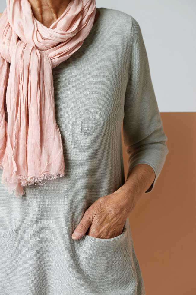 A woman wearing a pink scarf and grey sweater with her hand in the sweater's pocket. 
