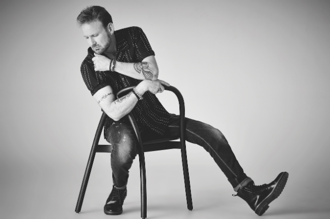 A picture of Corey Hart leaning back in a ming chair with his chin in his hand.