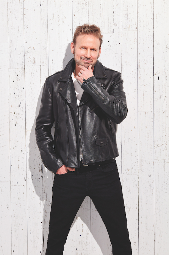 A photo of Corey Hart in a black biker jacket, standing against a white wooden fence.