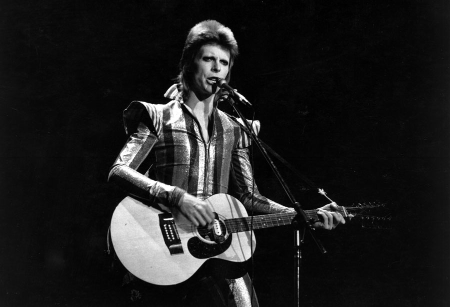 David Bowie as Ziggy Stardust on stage playing his guitar. 